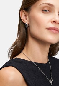 Zilveren oorbellen met een bladvormig ontwerp, gecombineerd met een ketting met een abstracte hanger. Het model draagt een zwarte textuurtop.
