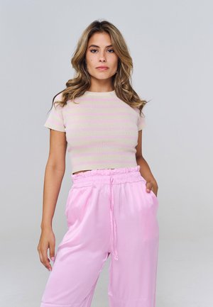 Donna con capelli biondi ondulati che indossa un crop top a righe rosa e beige a maniche corte e pantaloni rosa chiaro a vita alta, in piedi con una mano in tasca.