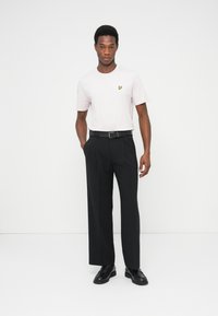 Hellrosa T-Shirt mit kleinem gelbem Logo, kombiniert mit schwarzer, maßgeschneiderter Hose und schwarzem Gürtel, abgerundet mit schwarzen Schuhen.