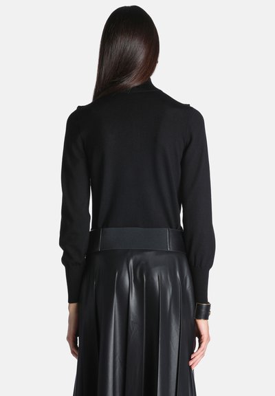 Femme aux longs cheveux foncés portant un haut noir à manches longues, une large ceinture et une jupe plissée en cuir noir, vue de dos.