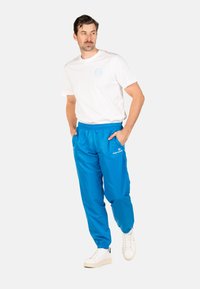 Weißes T-Shirt mit Rundhalsausschnitt, blaue Jogginghose mit elastischem Bund und seitlichen Taschen, sowie weiße Turnschuhe. Glattes Material; Logo auf der Jogginghose.