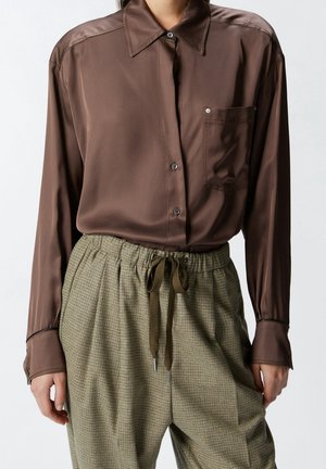 Personne portant une chemise marron brillante boutonnée avec une poche et un pantalon à carreaux beige à cordon visible du cou jusqu'à mi-cuisse.