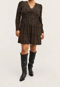 Femme portant une robe noire avec de petites impressions florales orange et des manches longues, accompagnée de bottes en cuir noires jusqu'aux genoux.