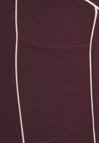 Primo piano di un tessuto bordeaux con piping bianco verticale e un dettaglio di cucitura orizzontale.