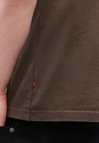 Κοντινή λήψη ενός ατόμου που φοράει καφέ πουκάμισο με μια μικ ρή κόκκινη ετικέτα Levi's στη ραφή της πλευράς και μαύρο παντελόνι με ορατό κουμπί.