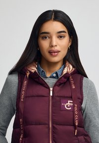 Burgundy pufferväst med en blänkande roséguld-fodring, med "POLO CLUB"-logotyp och dragkedjestängning. Bärs över en grå tröja.