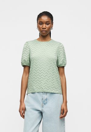 Kvinne med flettehår iført en lys grønn bluse med sikksakk-mønster og korte puffede ermer, samt lyseblå høytlivsjeans, stående foran ensfarget bakgrunn.