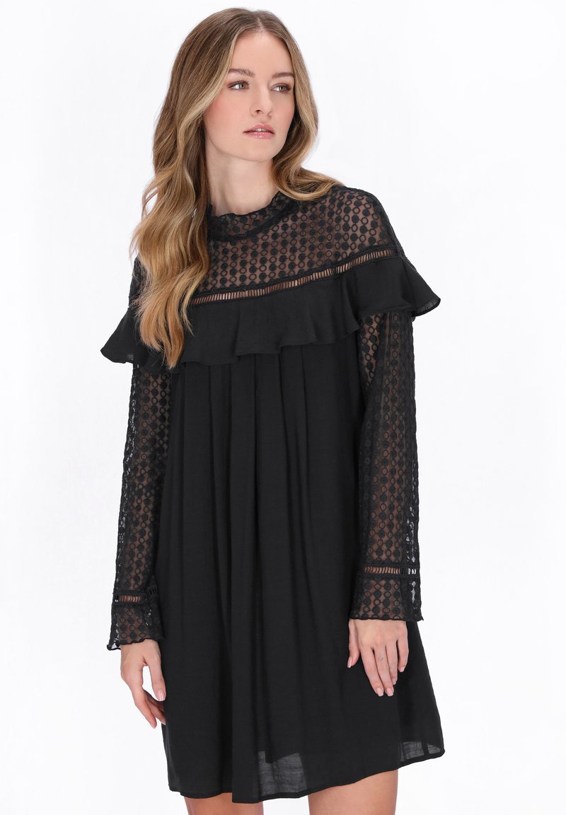 Hosszú, hullámos hajú nő fekete ruhában, áttetsző csipkés ujjakkal és felsőrésszel, amely fodros, vízszintes díszítéssel fut a mellkas fölött.