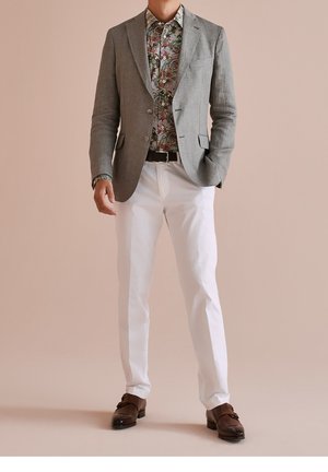 Grauer Blazer über einem mehrfarbigen Blumenshirt, kombiniert mit weißen Hosen und braunen Lederschuhen. Der Blazer hat einen strukturierten Stoff und einen Reverskragen.
