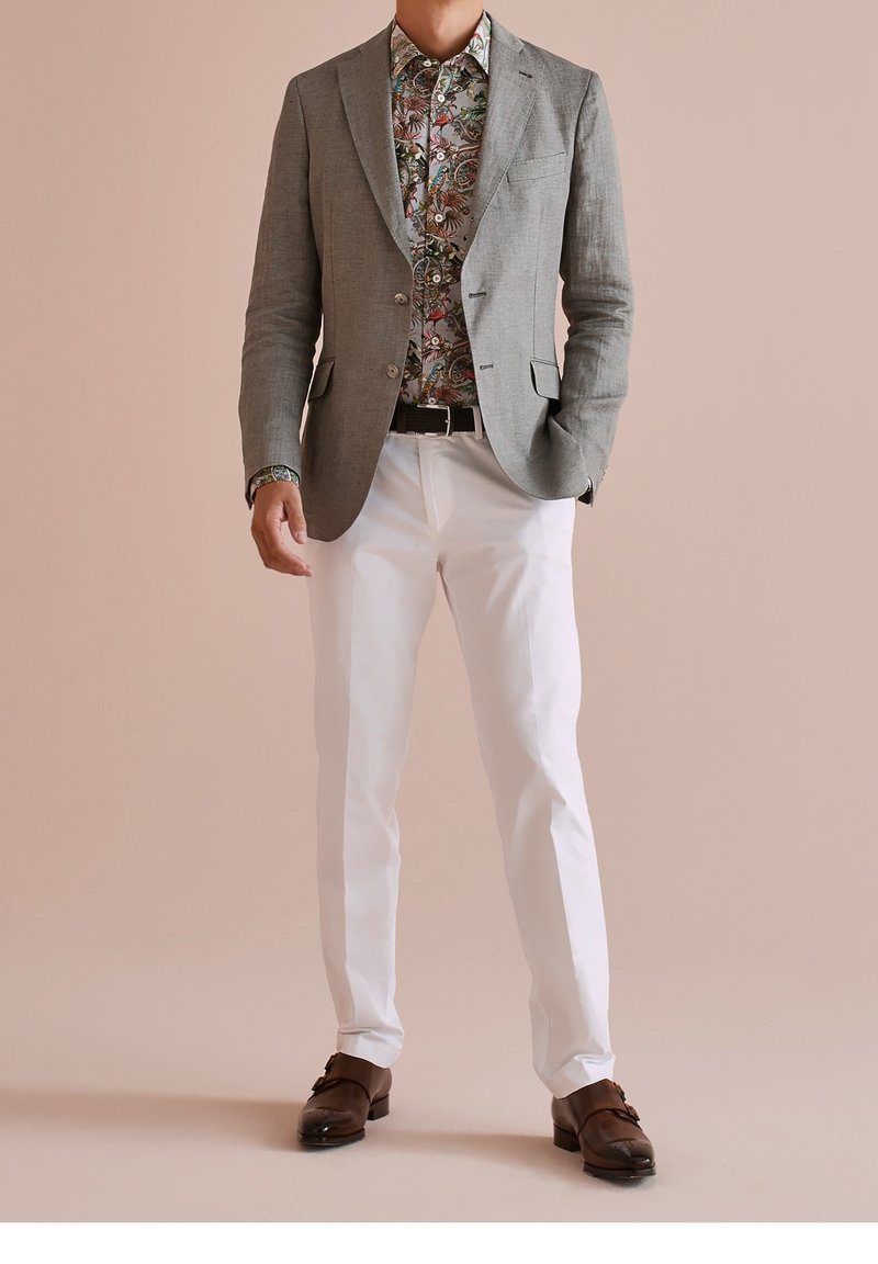 Grauer Blazer über einem mehrfarbigen Blumenshirt, kombiniert mit weißen Hosen und braunen Lederschuhen. Der Blazer hat einen strukturierten Stoff und einen Reverskragen.