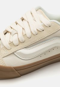 Beige suede sneaker with cream leather accents, flat laces, and a textured gum rubber sole; notable white stripe detail along the side.