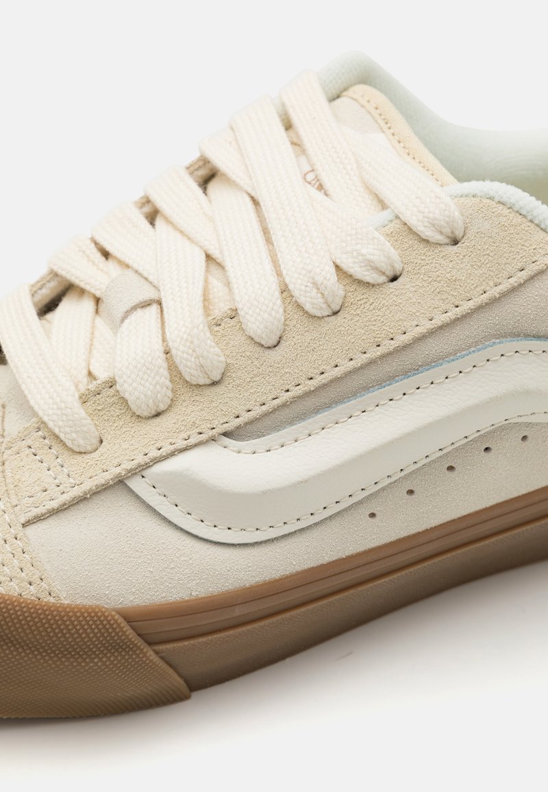 Zapatillas de ante beige con detalles de cuero crema, cordones planos y una suela de goma texturizada; destaca una línea blanca a lo largo del costado.