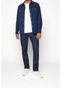 Camisa azul marino abotonada con dos bolsillos en el pecho, camiseta blanca de cuello redondo, vaqueros oscuros y zapatillas blancas, de pie frente a un fondo claro.