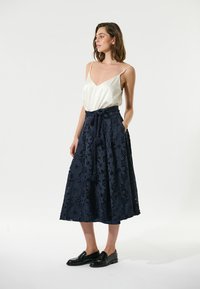 Gonna maxi in pizzo blu navy con motivo floreale, vita legata, texture a strati e tasche. Abbinata con un top in satin bianco e mocassini neri.