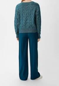 Person in einem türkisfarbenen, kabelgestrickten Pullover und lockeren, weit geschnittenen türkisfarbenen Hosen, der vor einem schlichten weißen Hintergrund steht und sich abwendet.