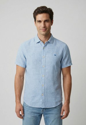 Mand iført en kortærmet, lyseblå og hvidstribet skjorte med knapper og krave, kombineret med lyseblå jeans, stående mod en ensfarvet baggrund.