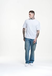 Camiseta blanca de manga corta, jeans holgados de color azul claro, zapatillas blancas, con tatuajes visibles en los brazos, de pie sobre un fondo sencillo.