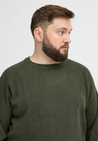 Pull en rib d'un vert olive avec un col rond et des manches longues. Texture douce et coupe décontractée, avec des détails de couture subtils.