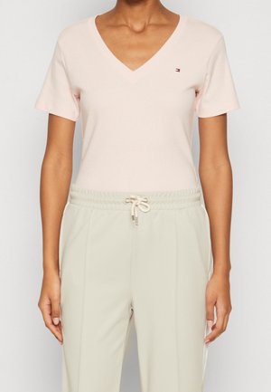 Femme portant un haut court à col en V rose clair avec un petit logo et un pantalon beige à cordon, debout devant un fond uni.