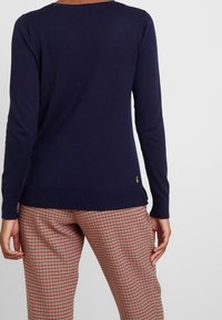 Marinblå stickad tröja med långa ärmar, ribbad nederkant och diskret logodetalj. Bärs med rutiga byxor i röd, svart och vit.