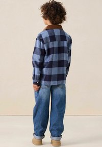 Chemise à carreaux bleu et marine avec un col en velours côtelé marron, portée avec un jean bleu ample et des chaussures beiges, vue de dos.