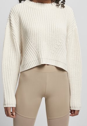 Woman wearing a cream chunky knit cropped sweater and beige high-waisted leggings against a plain background.