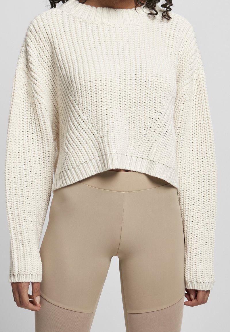 Femme portant un pull court en grosse maille crème et des leggings beige taille haute sur un fond uni.