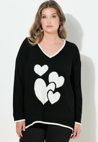Černý pletený svetr s výstřihem do V, zdobený bílými vzory ve tvaru srdcí. Kontrastní bílý lem podél výstřihu a spodního okraje. Volný střih.