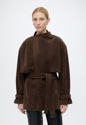 Frau mit blondem Haar, die goldene Creolen trägt und eine braune Wildlederjacke mit Gürtel, übergroßen Ärmeln und hohem Kragen trägt.