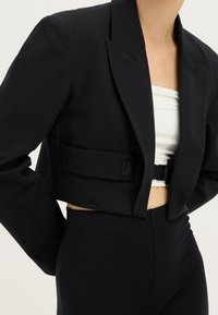 Kort svart kavaj med breda slag, bärs över en vit bandeau-top, ihop med höga ribbade svarta byxor.