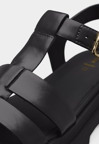 Μαύρο δερμάτινο σανδάλι με φαρδιές τιράντες, ρυθμιζόμενη αγκράφα σε χρυσαφί τόνο και επενδεδυμένη σόλα.