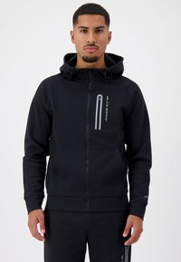 Zwarte rits hoodie van zachte stof met een hoge hals, twee voorzakken en reflecterende details. Model heeft handen in de zakken.