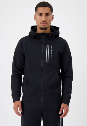 Black zip-up hoodie in soft fabric with a high neckline, two front pockets, and reflective detailing. Model has hands in pockets.