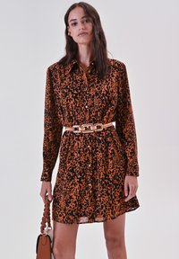 Robe chemise à imprimé léopard en orange et noir, avec un bouton de fermeture, des manches longues et une taille ceinturée. Accompagnée d'un sac à main marron.