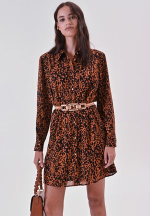 Leopardprint shirtjurk in oranje en zwart, uitgerust met een knoopsluiting, lange mouwen en een ceintuurlijn. Vergezeld door een bruine handtas.