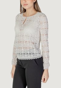 Top in pizzo a maniche lunghe con motivo floreale; colore grigio chiaro, scollatura a chiave e orlo a onde; indossato con pantaloni attillati scuri.