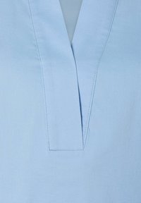 Helles blaues Blusenshirt mit V-Ausschnitt und sauberer Nahtverarbeitung. Der Stoff wirkt glatt und hat eine weiche Textur. Keine sichtbaren Verzierungen.