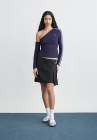 Νεαρή γυναίκα στέκεται σε μπλε χαλί, φορώντας μονομπατιέρα μωβ μπλούζα με μακρύ μανίκι, μαύρη φούστα με φερμουάρ στο μπροστινό μέρος, λευκές κάλτσες και ασημένια παπούτσια.