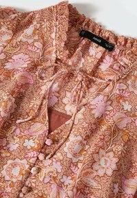 Blouse à motifs floraux avec des couleurs rose, orange et crème, présentant un col à volants, un col à nouer et des boutons recouverts de tissu sur le devant.