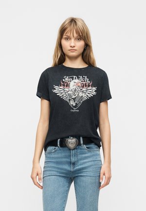 Noor naine kannab musta Von Dushi kujundusega t-särki, millel on kotkas ja leegid, siniseid pükse ning südamekujulise pandlaga vööd, seistes valgel taustal.