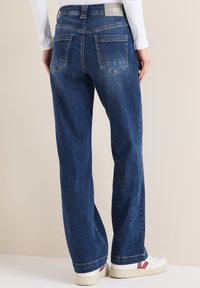 Tmavě modré džínové kalhoty s širokými nohavicemi a zadními kapsami, s pasem ve střední výšce a jemnými švovými detaily. Kombinovány s bílými teniskami.