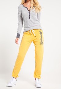 Pruhovaný dlouhý rukáv s šedými detaily a knoflíky, doplněný o jasně žluté tepláky se šedým logem. Celý vzhled doplňují bílé tenisky.