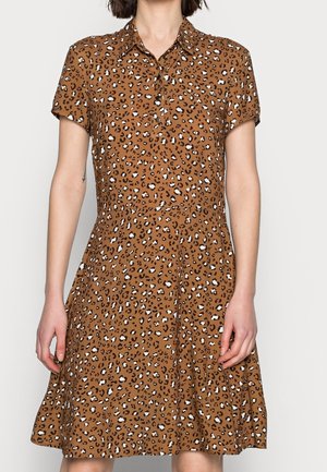 Donna che indossa un vestito marrone a maniche corte con colletto e stampa leopardata bianca e nera, in piedi su uno sfondo chiaro uniforme.
