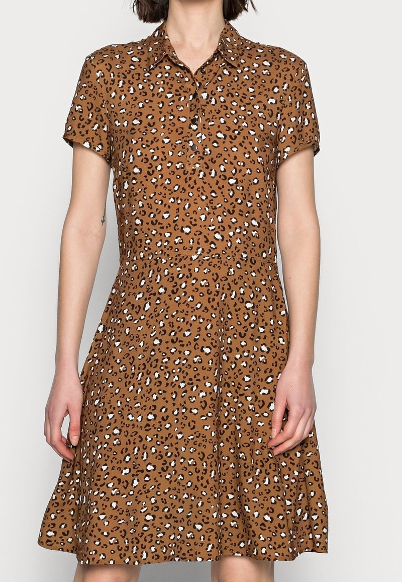 Femme portant une robe marron à manches courtes avec col, imprimé léopard noir et blanc, debout devant un fond clair uni.