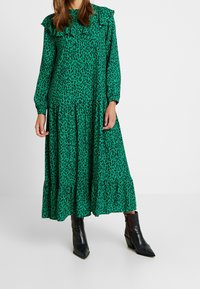 Groene maxi-jurk met zwarte luipaardvlekken, ruches aan de halslijn, lange mouwen en een gelaagde rok, gecombineerd met zwarte enkellaarsjes met hak.