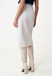 Falda midi de mezclilla blanca con una abertura trasera, que cuenta con dos bolsillos en la parte posterior. Combinada con botas de tacón de color beige claro, acentuando una silueta ajustada.