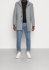 Man som bär en grå knappad kappa, svart stjärnmönstrad tröja, uppvikta blå jeans, vita strumpor och vita sneakers, stående inomhus.