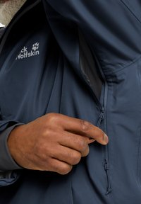 Giacca impermeabile blu navy con zip, lacci elastici e logo Jack Wolfskin ben visibile. Tessuto dalla texture liscia.
