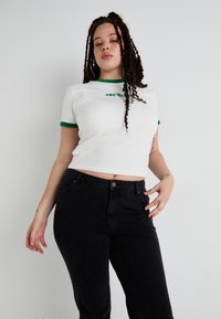 White cropped t-shirt with green accents and logo, paired with high-waisted black jeans. The outfit features a smooth texture and minimal design.