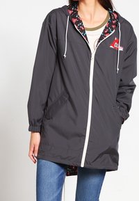 Czarna kurtka przeciwdeszczowa z kwiatowym podszewką kaptura, zamkiem błyskawicznym z przodu, dwiema kieszeniami i elastycznymi mankietami. Luźny krój idealny do warstwowego ubierania.